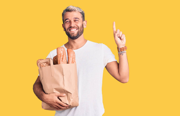 Fiatal, jóképű szőke férfi kezében papírzacskó kenyérrel meglepve egy ötlettel vagy kérdéssel ujjal mutogatva boldog arccal, első számú  - Fotó, kép