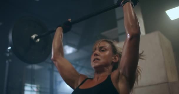 a female weightlifter performs a barbell lift in a dark gym. a woman lifting a heavy bar over her head - Filmati, video