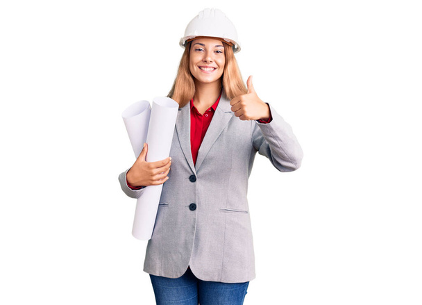 Piękna młoda kobieta nosi architekt twardy kapelusz trzymając budować projekt uśmiechnięty szczęśliwy i pozytywny, kciuk do góry robi doskonałe i znak aprobaty  - Zdjęcie, obraz