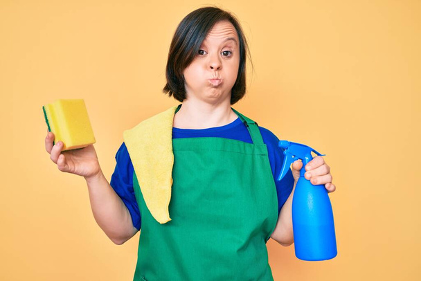 Brunette woman with down syndrome wearing apron holding scourer puffing cheeks with funny face. mouth inflated with air, catching air.  - Photo, Image