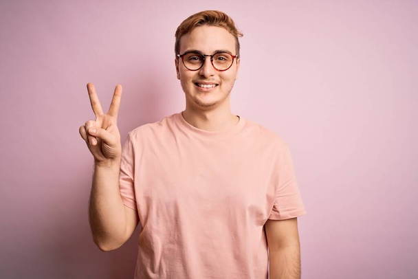 Joven hombre pelirrojo guapo con camiseta casual de pie sobre un fondo rosa aislado sonriendo mirando a la cámara mostrando los dedos haciendo señal de victoria. Número dos.
. - Foto, imagen