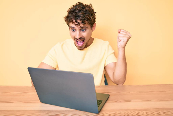 Młody biały mężczyzna z kręconymi włosami pracuje w biurze z laptopem krzycząc dumny, świętując zwycięstwo i sukces bardzo podekscytowany podniesionymi ramionami  - Zdjęcie, obraz