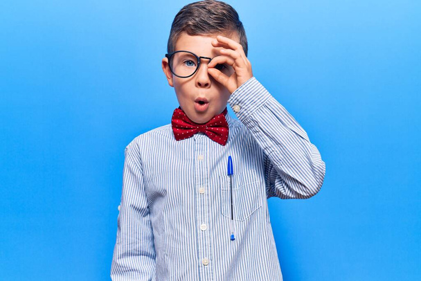 Lindo niño rubio con corbata de lazo nerd y gafas haciendo buen gesto conmocionado con la cara sorprendida, los ojos mirando a través de los dedos. expresión incrédula.  - Foto, imagen