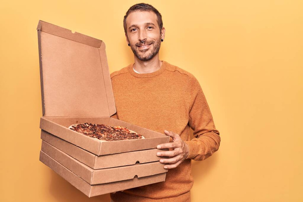 Młody przystojny mężczyzna trzyma pudełka po pizzy patrząc pozytywnie i szczęśliwy stojąc i uśmiechając się z pewnym uśmiechem pokazując zęby  - Zdjęcie, obraz