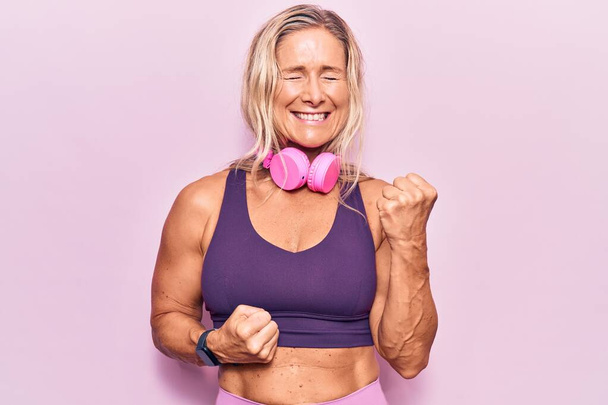 Middle age caucasian blonde woman wearing gym clothes and using headphones celebrating surprised and amazed for success with arms raised and eyes closed  - Photo, Image