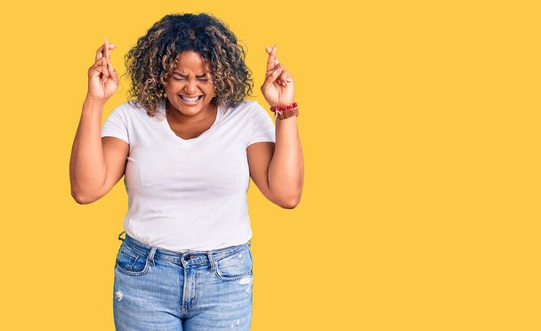 Młoda Afroamerykanka plus rozmiar kobieta w luźnych ubraniach gestykulujący palec skrzyżowany uśmiechnięty z nadzieją i zamkniętymi oczami. szczęście i przesądna koncepcja.  - Zdjęcie, obraz