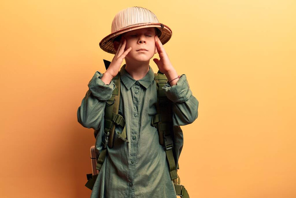 Cute blond kid wearing explorer hat and backpack with hand on head, headache because stress. suffering migraine.  - Photo, image