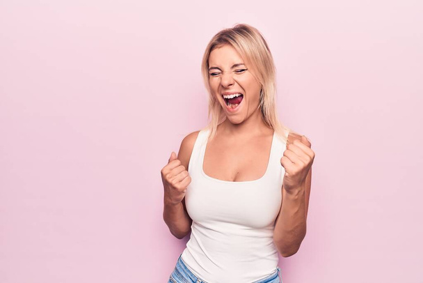 Junge schöne blonde Frau in lässigem T-Shirt steht vor isoliertem rosa Hintergrund und feiert überrascht und erstaunt über den Erfolg mit erhobenen Armen und geschlossenen Augen - Foto, Bild