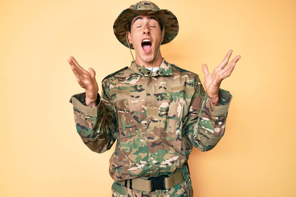 Jeune bel homme portant un uniforme de camouflage de l'armée célébrant fou et fou de succès avec les bras levés et les yeux fermés hurlant excité. concept gagnant  - Photo, image
