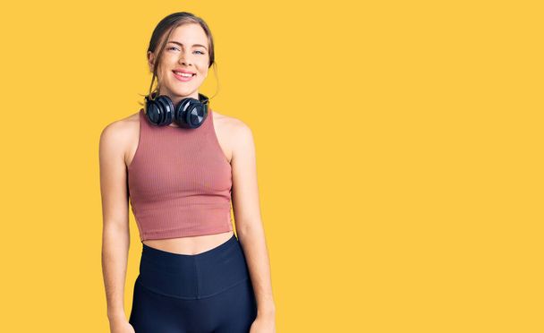 Beautiful caucasian young woman wearing gym clothes and using headphones with a happy and cool smile on face. lucky person.  - 写真・画像