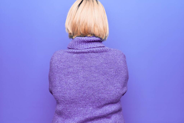 Hermosa mujer rubia de talla grande con suéter casual de cuello alto sobre fondo púrpura mirando hacia atrás con los brazos cruzados
 - Foto, imagen
