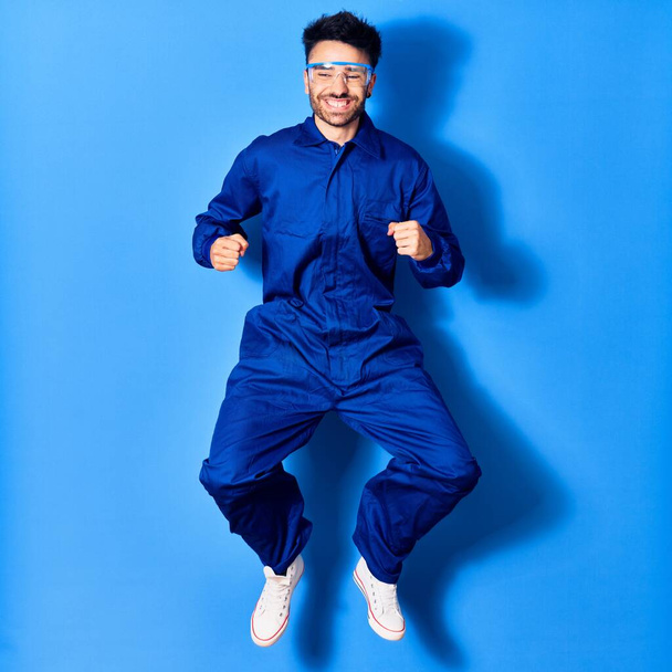 Young handsome hispanic man wearing painter uniform and glasses smiling happy. Jumping with smile on face doig winner sign with fists up over isolated blue background - Photo, Image