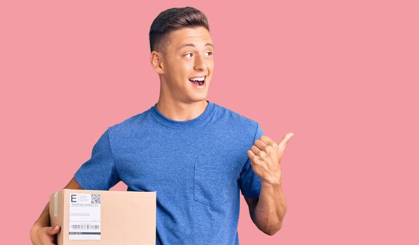 Young handsome hispanic man holding delivery package pointing thumb up to the side smiling happy with open mouth  - Photo, Image
