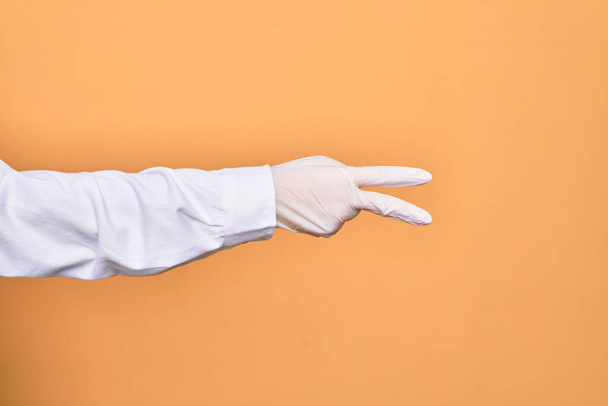 Hand of a person wearing medical surgical glove showing two fingers doing victory sign over isolated yellow background - Foto, Imagen