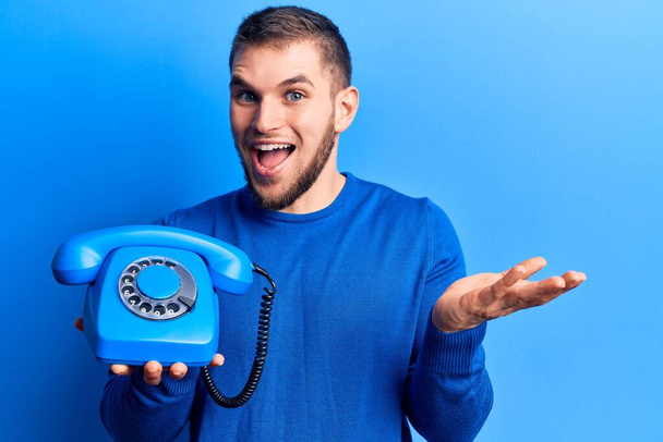 Jovem homem bonito segurando telefone vintage celebrando realização com sorriso feliz e expressão vencedora com mão levantada
  - Foto, Imagem