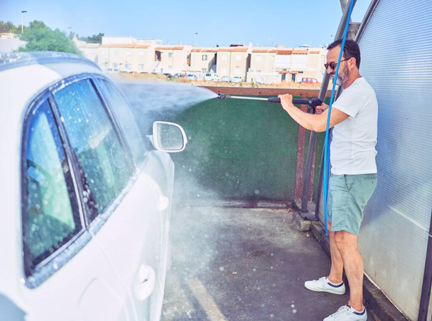 Middle age handsome man wearing casual clothes and sunglasses smiling happy. Standing with smile on face washing car using water pistol. - Photo, Image