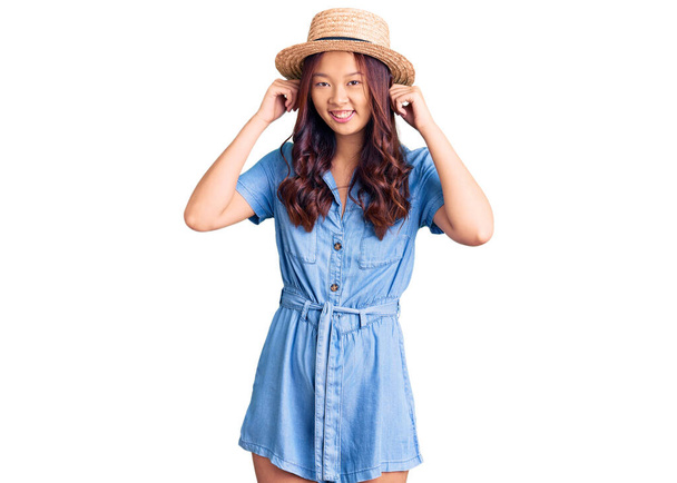 Joven hermosa chica china con sombrero de verano sonriendo tirando de las orejas con los dedos, gesto divertido. problema de audición
  - Foto, Imagen