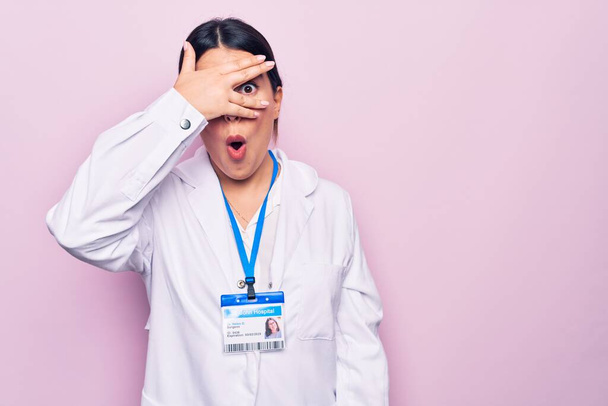 Young beautiful doctor woman wearing stethoscope and id card over isolated pink background peeking in shock covering face and eyes with hand, looking through fingers afraid - Zdjęcie, obraz