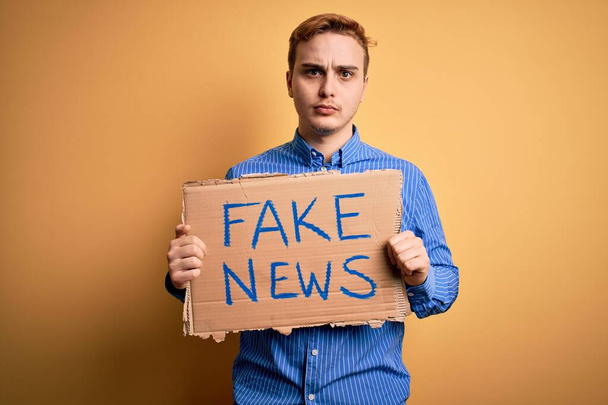 Junger gutaussehender rothaariger Mann mit Transparent mit Fake-News-Botschaft über gelbem Hintergrund und nüchternem Gesichtsausdruck, der selbstbewusst wirkt - Foto, Bild