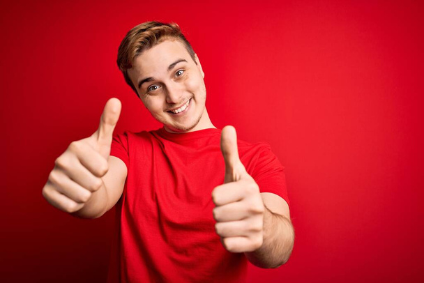 Giovane bel rossa uomo indossa casual t-shirt su sfondo rosso isolato approvando facendo gesto positivo con la mano, pollici in su sorridente e felice per il successo. Gesto del vincitore. - Foto, immagini