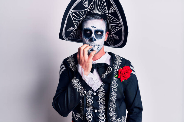 Молодой человек, одетый в мексиканский день мертвого костюма поверх белого, выглядящий напряженным и нервным с руками во рту, грызущими ногти. тревожные проблемы.  - Фото, изображение