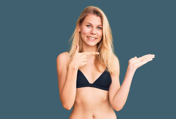 Junge schöne blonde Frau im Bikini überrascht und lächelt in die Kamera, während sie mit der Hand präsentiert und mit dem Finger zeigt.  - Foto, Bild