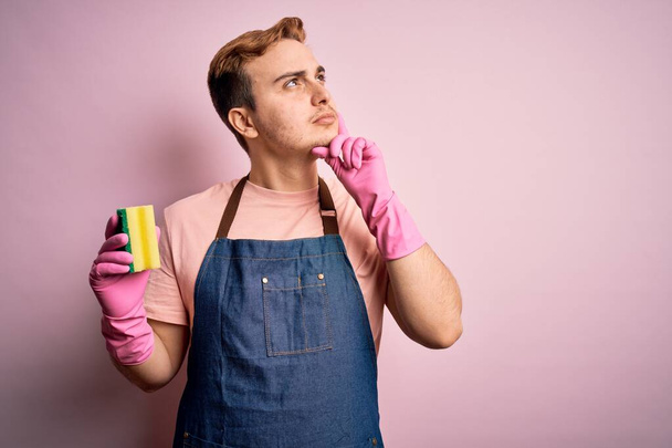 bel homme rousse faisant des travaux ménagers portant tablier et gants en utilisant nettoyeur visage sérieux réfléchissant à la question avec la main sur le menton, réfléchissant à l'idée déroutante - Photo, image