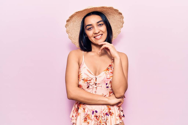 Yaz şapkası takan genç ve güzel bir Latin kadın kameraya kollarını kavuşturup çenesine dokunarak gülümsüyor. Olumlu düşünmek.  - Fotoğraf, Görsel