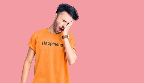 Young hispanic man wearing t shirt with happiness word message yawning tired covering half face, eye and mouth with hand. face hurts in pain.  - Photo, Image