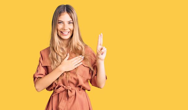 Beautiful caucasian woman with blonde hair wearing summer jumpsuit smiling swearing with hand on chest and fingers up, making a loyalty promise oath  - Photo, Image