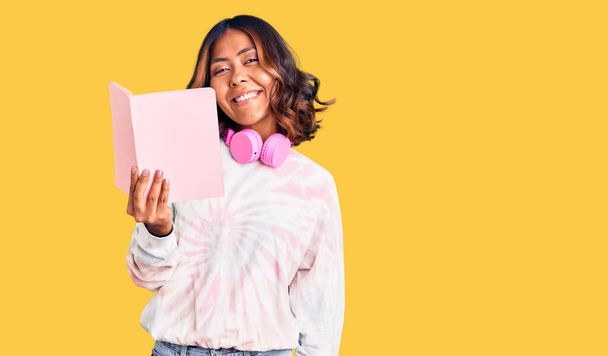 Young beautiful mixed race woman reading a book wearing headphones looking positive and happy standing and smiling with a confident smile showing teeth  - Foto, imagen