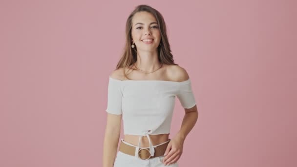 A beautiful smiling young woman is waving her hand to the camera isolated over pink background in studio - Materiał filmowy, wideo