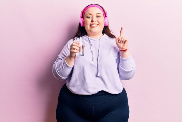 Young plus size woman drinking glass of water using headphones surprised with an idea or question pointing finger with happy face, number one  - Fotografie, Obrázek