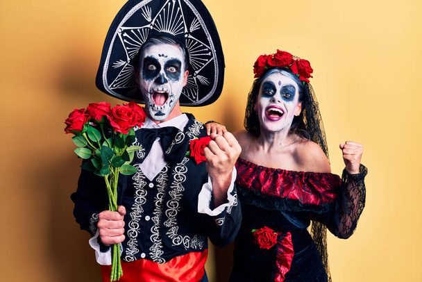 Young couple wearing mexican day of the dead costume holding roses screaming proud, celebrating victory and success very excited with raised arms  - Photo, Image