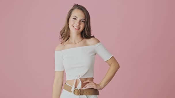 An attractive young woman is showing peace gesture to the camera standing isolated over pink background in studio - Imágenes, Vídeo