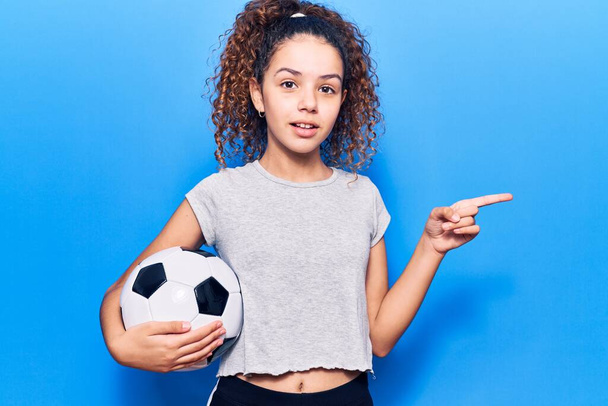Piękne dziecko dziewczyna z kręconymi włosami trzymając piłkę nożną uśmiechając szczęśliwy wskazując z ręki i palca na bok  - Zdjęcie, obraz