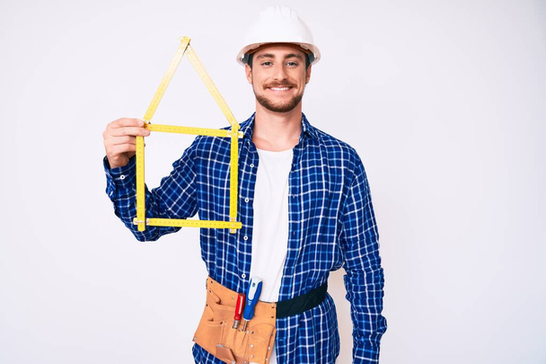 Mimar şapkalı yakışıklı genç adam inşaat projesini tutuyor. Pozitif ve mutlu görünüyor. Kendine güvenen bir gülümsemeyle dişlerini gösteriyor.  - Fotoğraf, Görsel