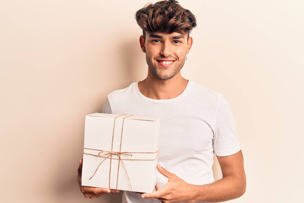 Junger hispanischer Mann mit Geschenk sieht positiv und glücklich stehend aus und lächelt mit einem selbstbewussten Lächeln, das Zähne zeigt  - Foto, Bild