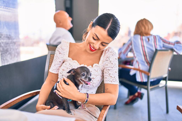 Jonge mooie brunette vrouw glimlachend gelukkig en zelfverzekerd. Zittend met een glimlach op het gezicht met chihuahua hond in restaurant - Foto, afbeelding