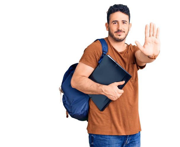 Jeune homme hispanique portant un sac à dos étudiant tenant un cartable avec une main ouverte faisant signe d'arrêt avec une expression sérieuse et confiante, geste de défense  - Photo, image