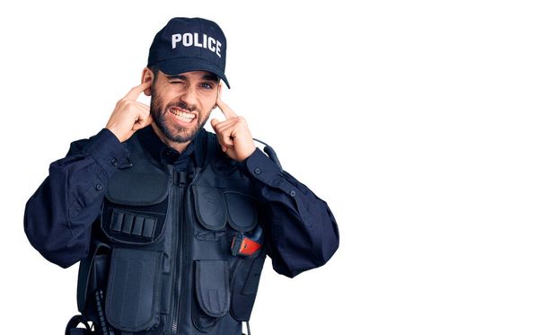 大きな音楽のノイズのためにいらいらする表情で指で耳を覆う警察の制服を着て髭を生やした若いハンサムな男。聴覚障害の概念.  - 写真・画像