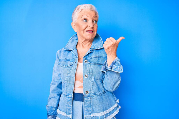 Ältere schöne Frau mit blauen Augen und grauen Haaren trägt lässigen Jeansstil lächelnd mit glücklichem Gesicht und zeigt mit erhobenem Daumen zur Seite.  - Foto, Bild