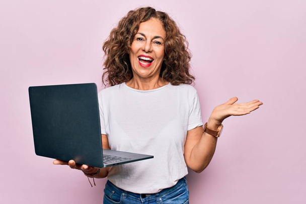 Middelbare leeftijd mooie zakenvrouw werken met behulp van laptop over geïsoleerde roze achtergrond vieren prestatie met vrolijke glimlach en winnaar uitdrukking met opgeheven hand - Foto, afbeelding