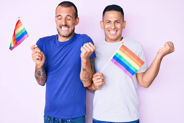 Νέοι γκέι ζευγάρι των δύο ανδρών που κρατούν σημαίες ουράνιο τόξο Igbtq μαζί ουρλιάζοντας υπερήφανοι, γιορτάζει τη νίκη και την επιτυχία πολύ ενθουσιασμένος με σήκωσε το χέρι  - Φωτογραφία, εικόνα