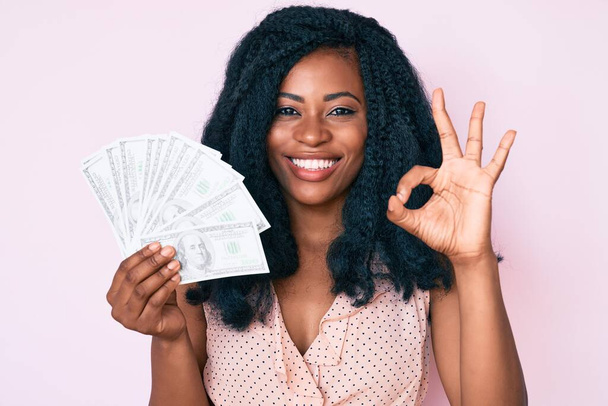 Dolarları tutan güzel Afrikalı kadın parmaklarıyla imza atıyor, gülümseyerek mükemmel bir sembol oluşturuyor.  - Fotoğraf, Görsel