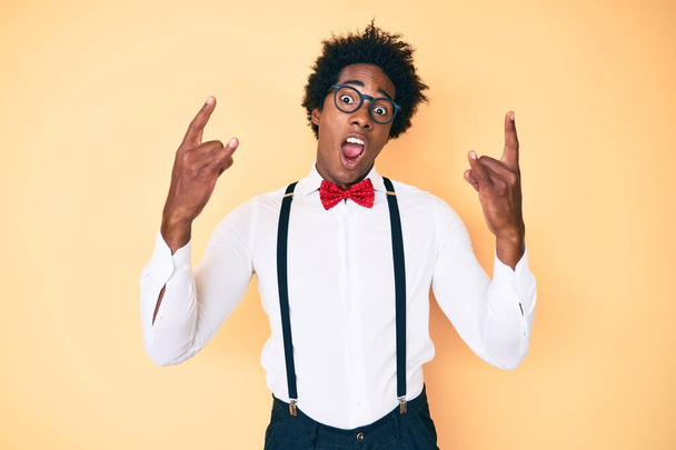 Schöner afrikanisch-amerikanischer Mann mit Afro-Haaren im Hipster-eleganten Look, der mit verrücktem Gesichtsausdruck Rock-Symbol mit erhobenen Händen macht. Musikstar. Schweres Konzept.  - Foto, Bild