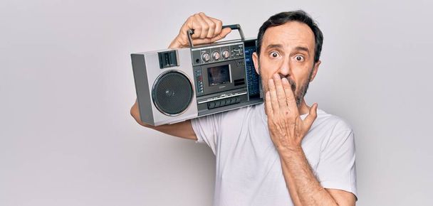 Middle age handsome man listening vintage boombox over isolated white background covering mouth with hand, shocked and afraid for mistake. Surprised expression - Photo, Image