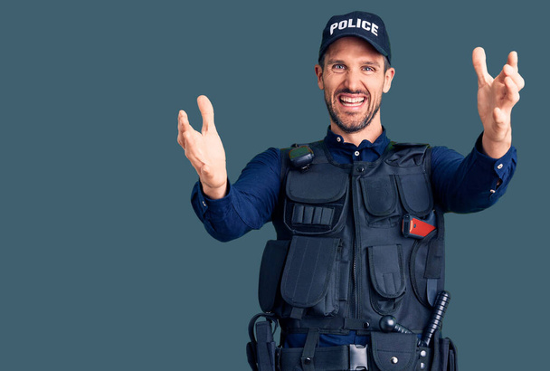 Joven hombre guapo con uniforme de policía mirando a la cámara sonriendo con los brazos abiertos para abrazar. expresión alegre abrazando la felicidad.  - Foto, imagen