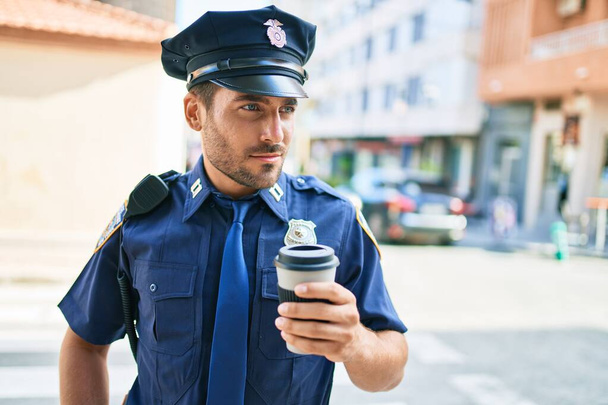 Genç İspanyol polisi ciddi bir ifadeyle polis üniforması giyiyor. Kasaba caddesinde dikilmiş kahve içerken. - Fotoğraf, Görsel