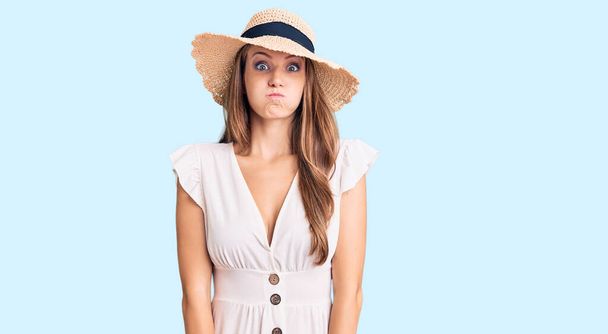 Young beautiful blonde woman wearing summer dress and hat puffing cheeks with funny face. mouth inflated with air, crazy expression.  - Фото, зображення